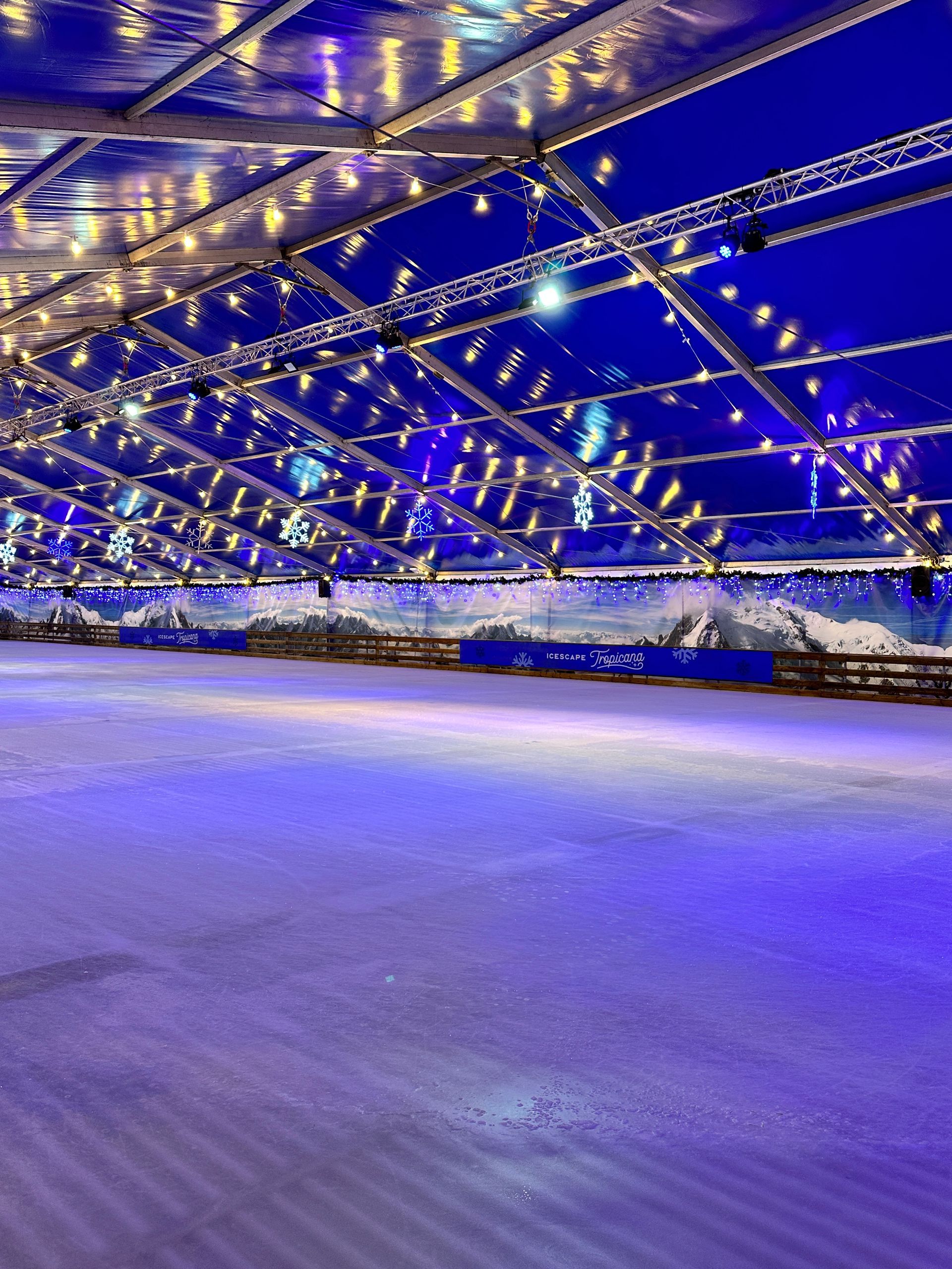 A large ice rink is lit up at night with christmas lights hanging from the ceiling.