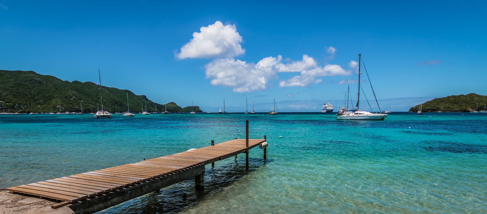 A wooden pier extends into turquoise water, with sailboats and green islands under a blue sky