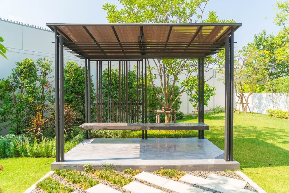Gazebo with dark metal frame and wooden benches in a green yard with a concrete floor and garden path.