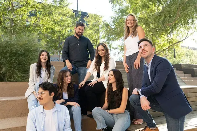 Group of six people posing outdoors on steps in a sunny park, smiling at the camera.
