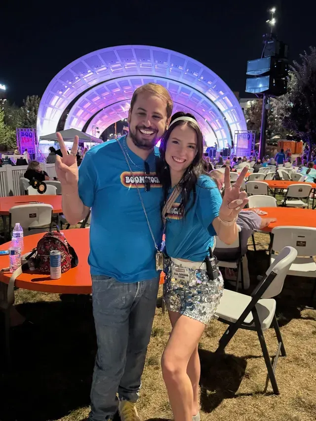 Couple posing at an outdoor concert, smiling in blue shirts before a lit stage at night