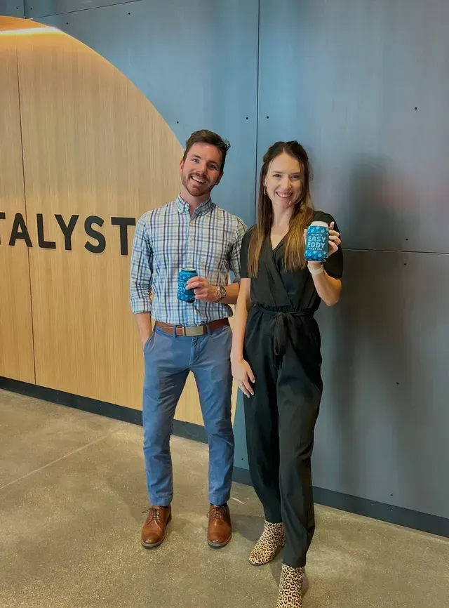 Two people posing indoors, smiling and holding blue cans in front of a wall with wood paneling.