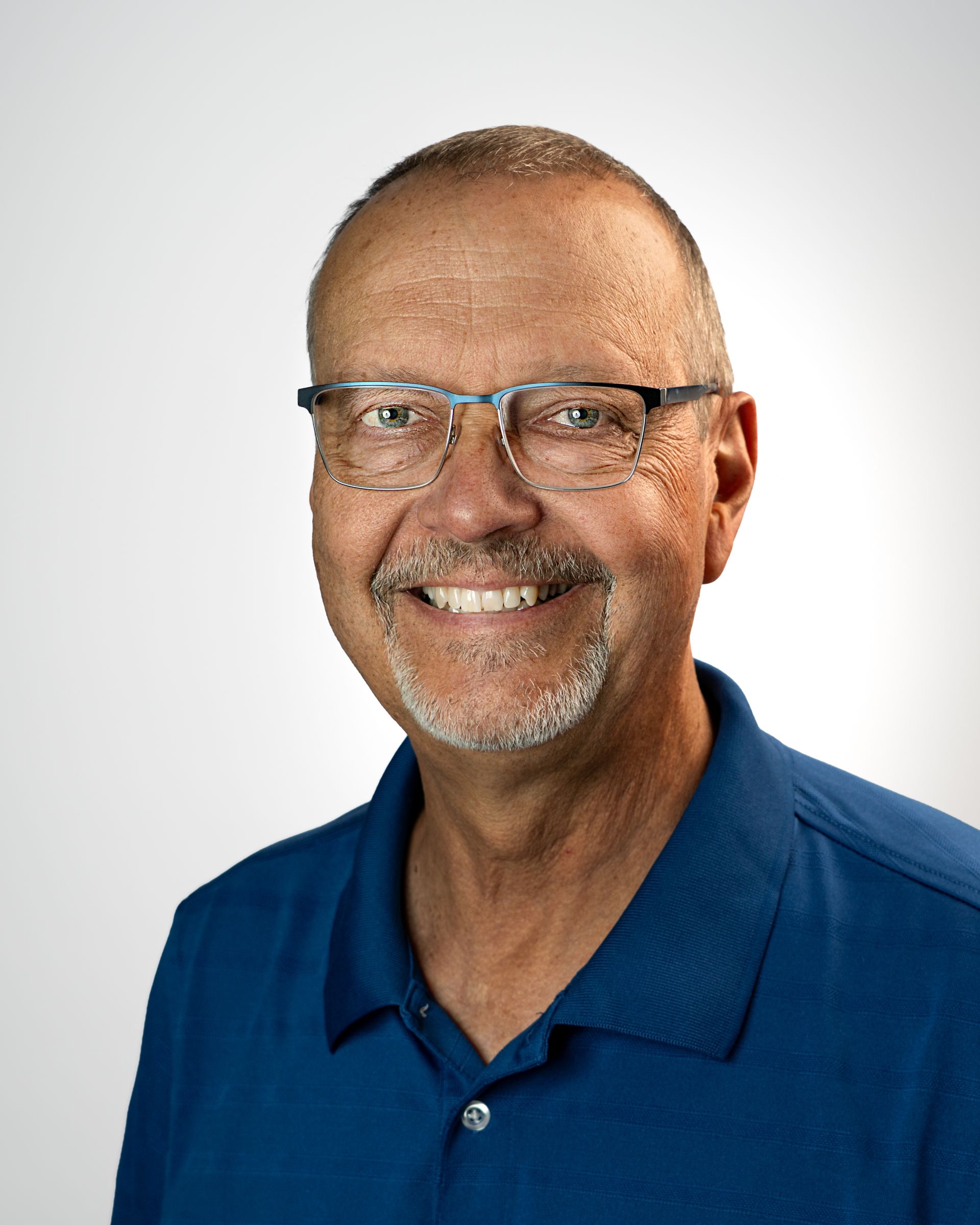 Smiling man in glasses and white pullover against a gray backdrop, wearing a logo on his chest.