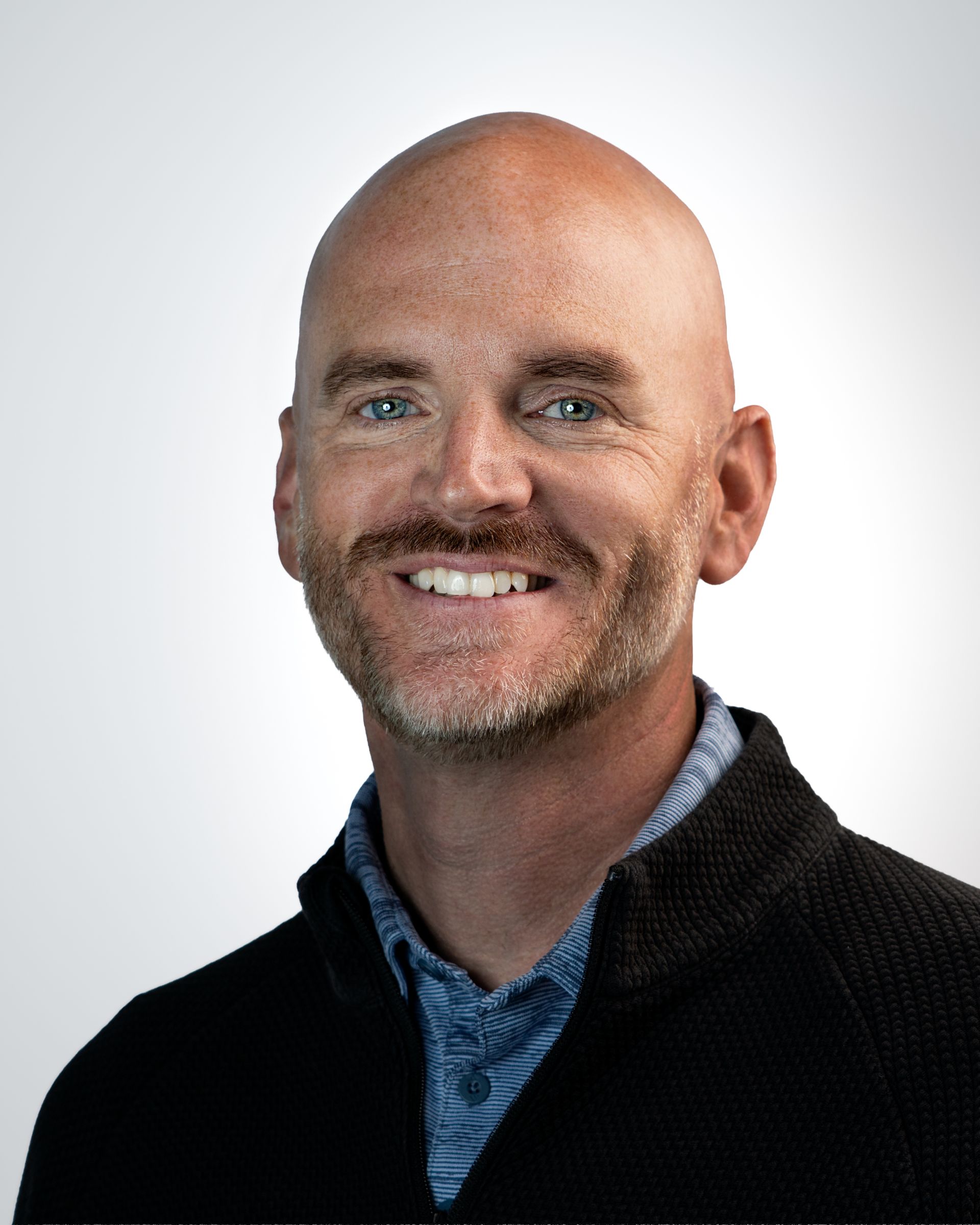 Professional headshot of a smiling bald man in a blue blazer and light blue shirt against a gray backdrop