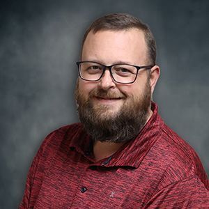 Smiling man with glasses and beard in a red shirt against a gray studio backdrop