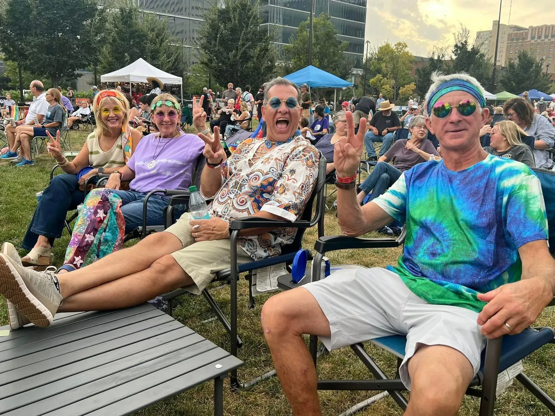 People relaxing in lawn chairs at an outdoor event, smiling and waving on a sunny day.