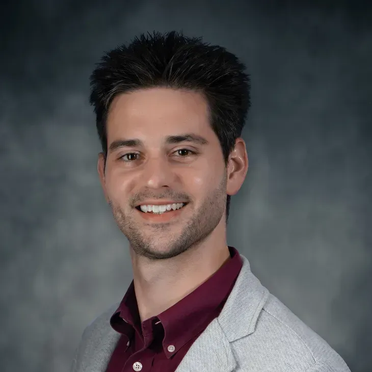 Studio portrait of a smiling man in a gray blazer and maroon shirt on a gray background
