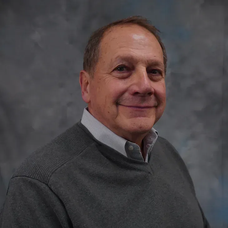 Smiling man in a gray sweater and collared shirt against a gray studio backdrop