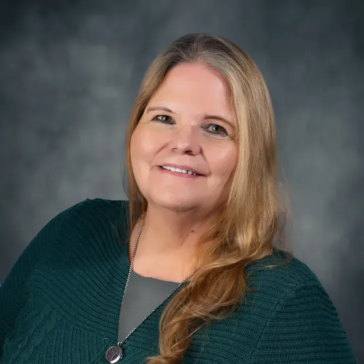 Smiling woman with long blonde hair wearing a dark green sweater against a gray studio backdrop