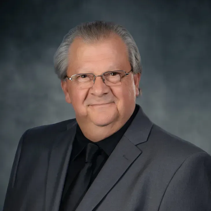 Studio portrait of a man in a gray suit and black shirt, wearing glasses, against a blue-gray backdrop