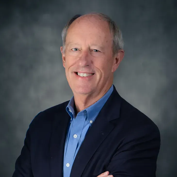 Portrait of a smiling man in a navy blazer and blue shirt against a gray studio background