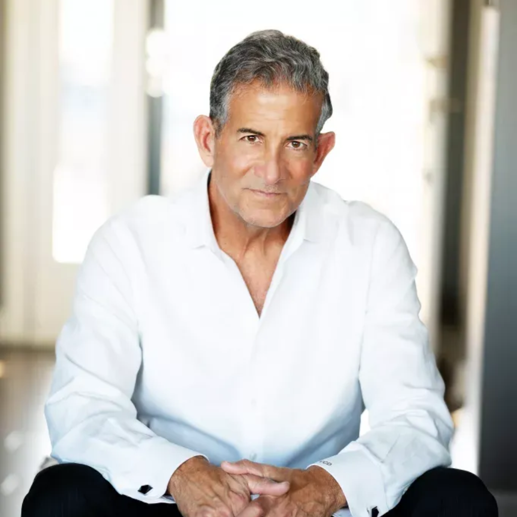 Man in white shirt seated indoors, facing forward with hands clasped