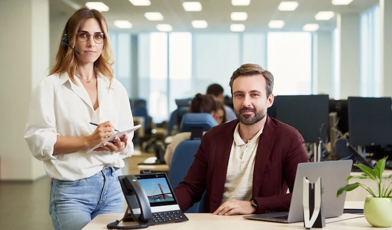 Donna con cuffie e uomo alla scrivania in ufficio. Donna che prende appunti, uomo al computer portatile, sorridenti.