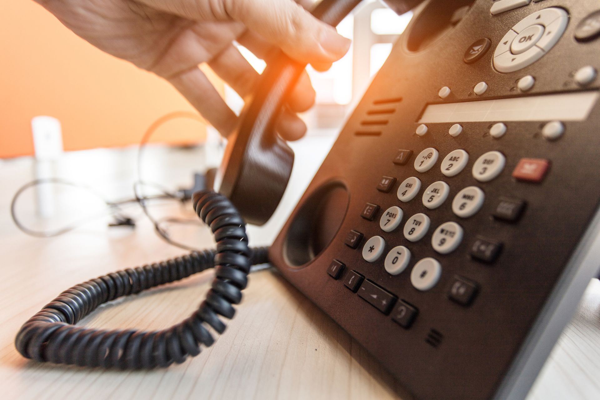 Mano che solleva un ricevitore telefonico nero, ambiente d'ufficio con scrivania e parete arancione.