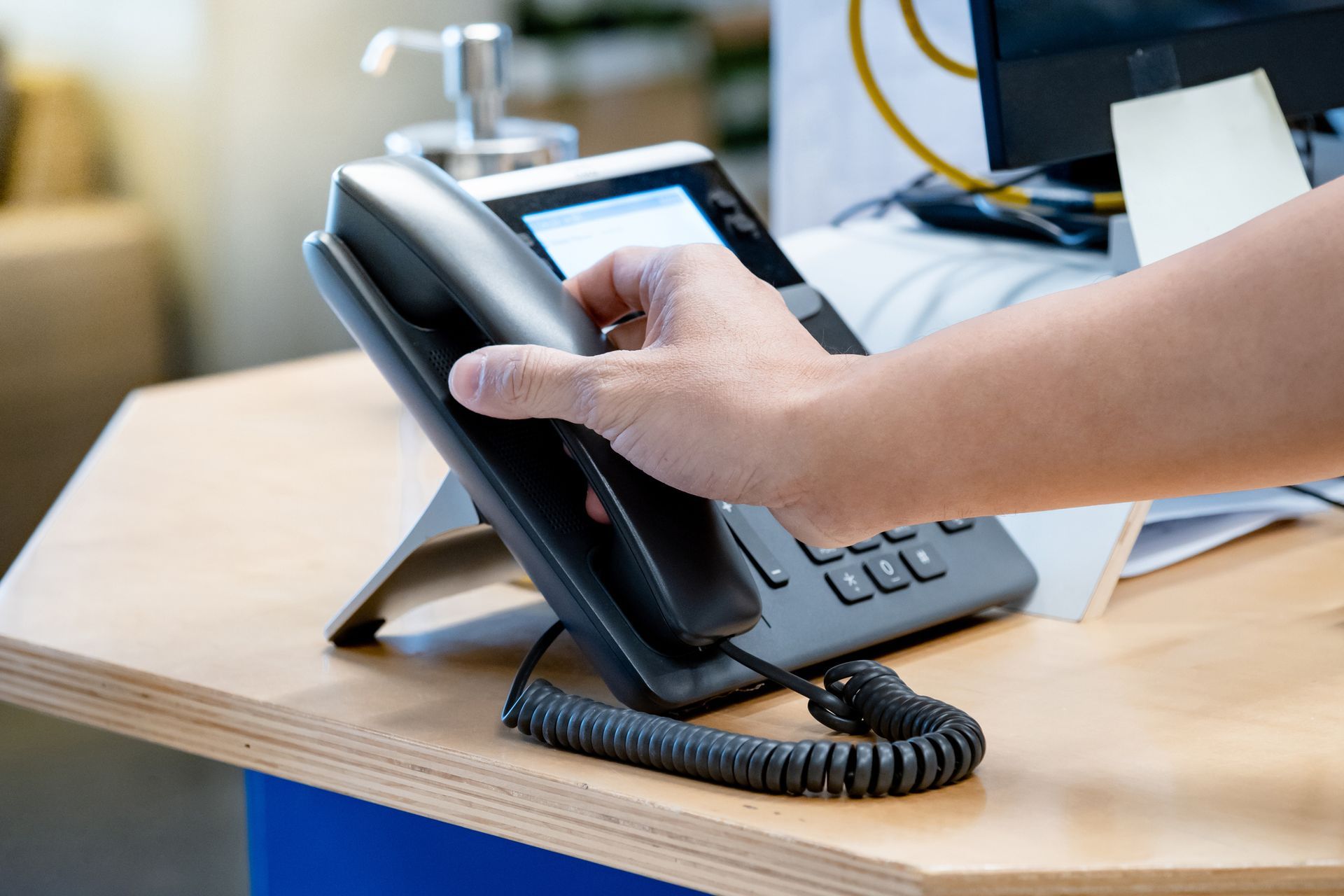Mano di una persona che allunga la mano verso un telefono nero da ufficio su una scrivania di legno. Il cavo del telefono è visibile.