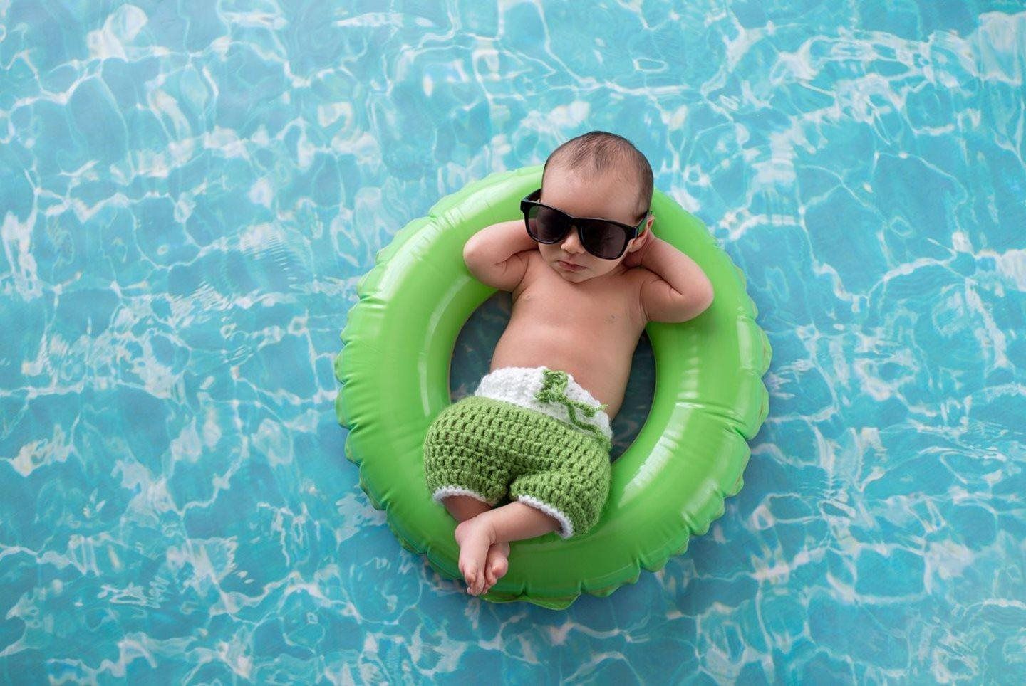 Baby floating on a green swim ring in a blue pool, wearing sunglasses and a knitted cover-up.