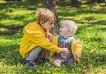 Two children in yellow jackets sitting on grass, one gently touching the other’s face.