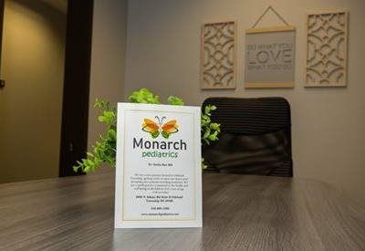 A Monarch Pediatrics information sign stands on a wooden desk, with wall art and a chair in the blurred background.