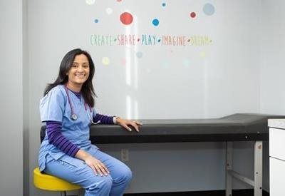 A professional in blue scrubs sits on a yellow stool in an exam room, smiling before a wall with colorful word decals.