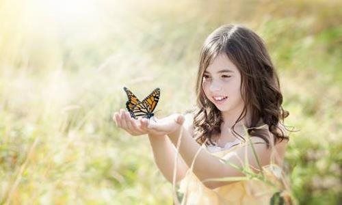 Young girl in a sunlit field gently holding a butterfly on her hand