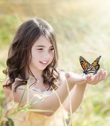 A person with long brown hair smiles gently, holding their open hands out as a monarch butterfly rests on their palm.