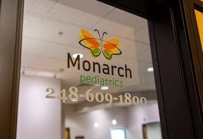A glass door displays the Monarch Pediatrics logo, featuring an orange and green butterfly and the phone number 248-609-1800.