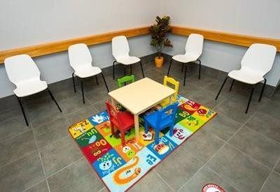 A small children's activity area with a colorful rug, a table, and chairs, surrounded by white chairs in a waiting room.