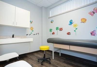 A pediatric exam room with white cabinets, a sink, a yellow stool, and a medical table against a wall with nature decals.