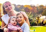 Woman and child smiling in a sunny park, holding a baby in pink.