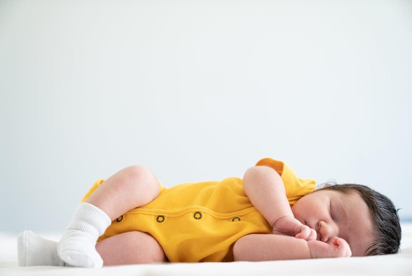 A baby wearing a yellow onesie and white socks sleeping on a soft white surface against a plain light background.