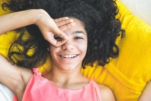 A person with curly dark hair wearing a pink top, lying on a yellow pillow and holding one hand in a circle over one eye.