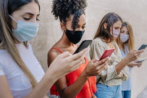 Four people wearing masks stand outdoors, looking at smartphones, with one in a red top holding a phone.