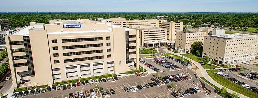An aerial view of the beige Beaumont Hospital complex with parking lots and surrounding trees under a clear blue sky.