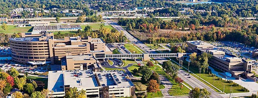 Aerial view of a large hospital campus with multiple tan buildings connected by walkways, surrounded by trees and roads.