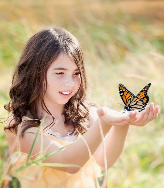 A person with long brown hair smiles gently, holding their open hands out as a monarch butterfly rests on their palm.