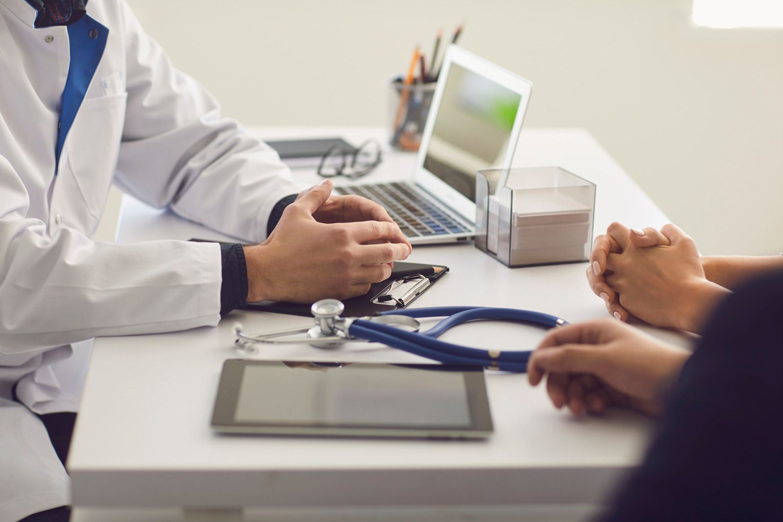 Doctor and patient at a desk, doctor gesturing, stethoscope and tablet on desk, laptop in background.