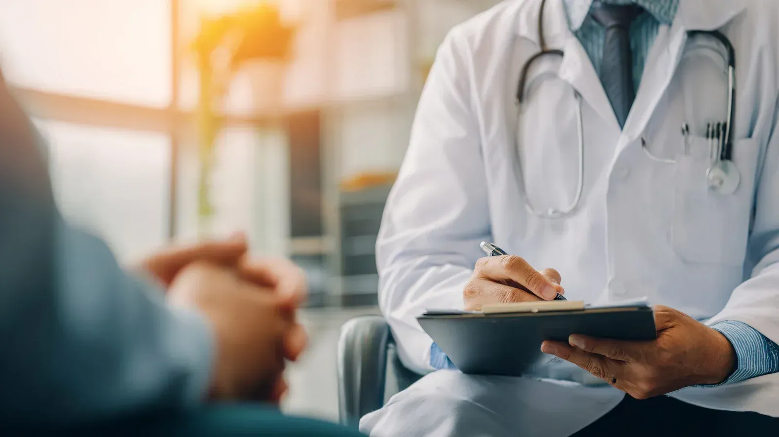 Doctor in white coat writing on clipboard, consulting with a patient in a bright office.