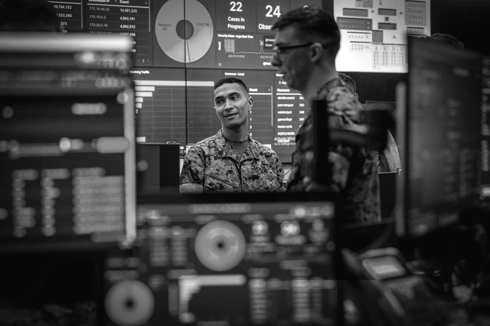Two military personnel in a control room with monitors displaying data and graphs. One is smiling.