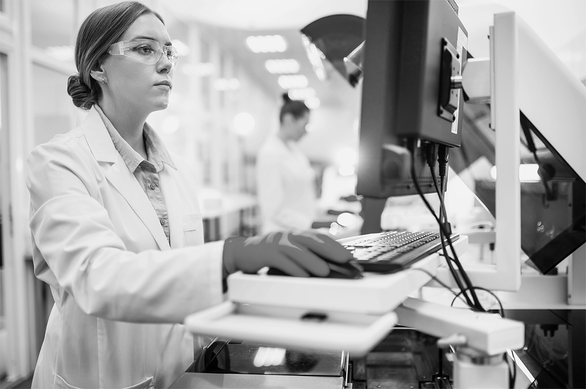 Scientist in lab coat and safety glasses uses computer, another person in the background.
