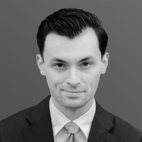 A man in a suit and tie, smiling slightly, with short, dark hair. Gray background.