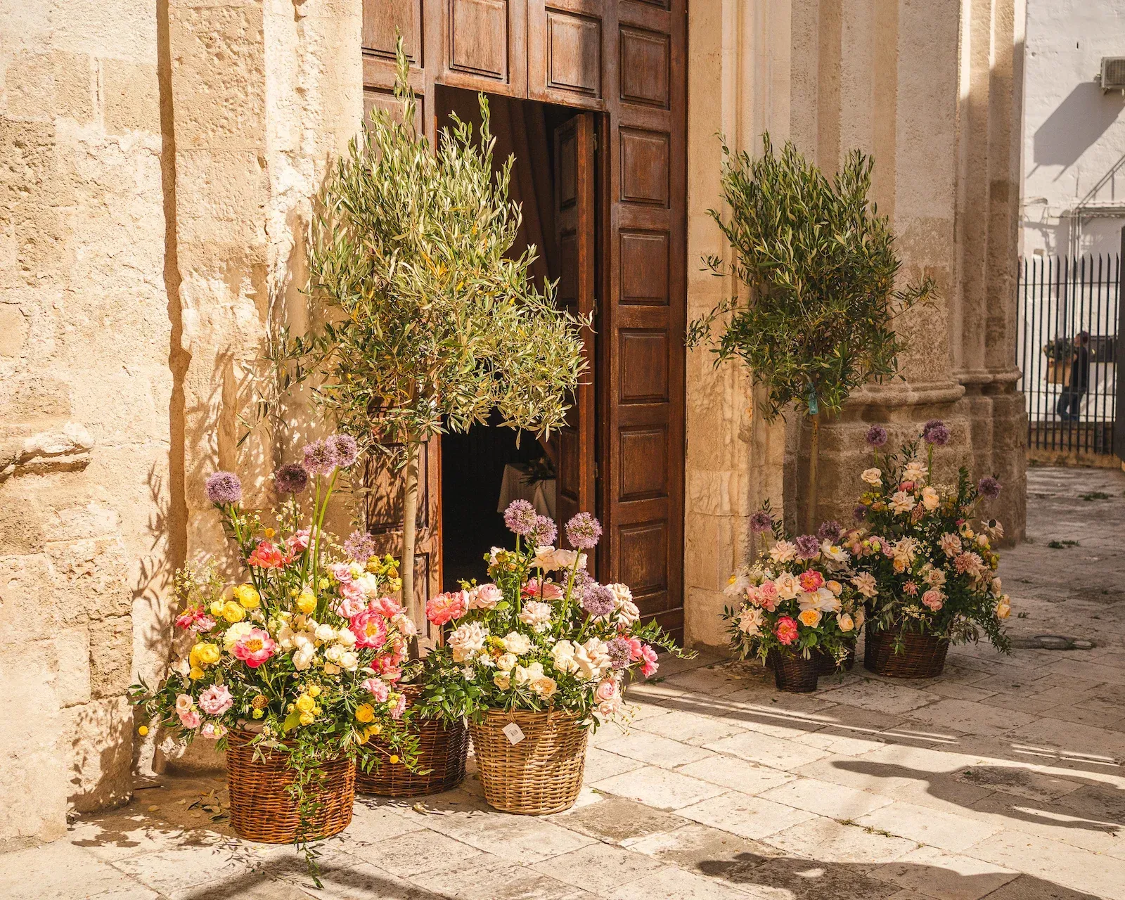 Ulivi e ceste di fiori ai lati del portale in legno di una chiesa in pietra