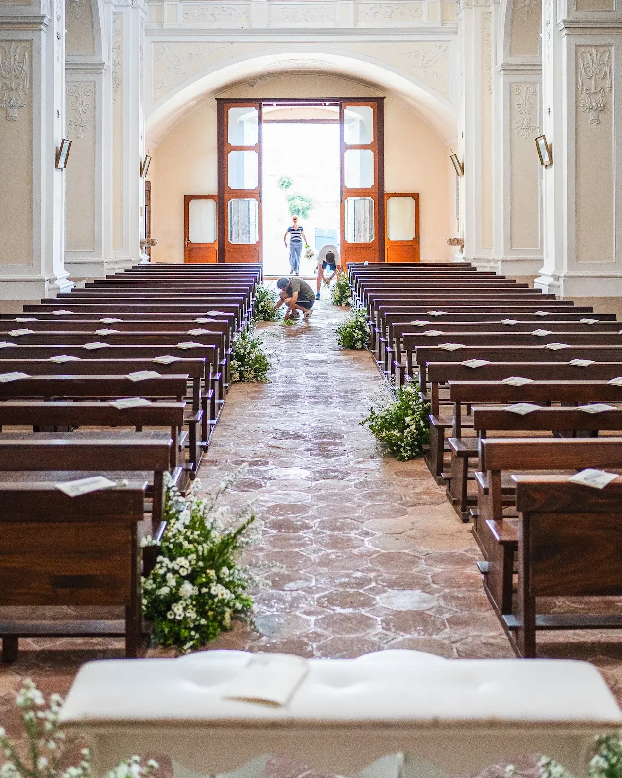Navata di una chiesa con i banchi in legno, mentre la squadra sistema i fiori bianchi lungo il corridoio
