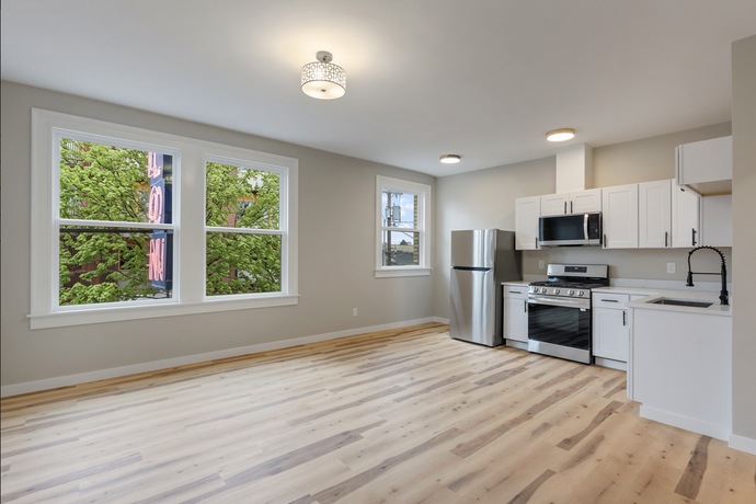 Empty modern kitchen with light wood floors, white cabinets, stainless steel appliances, and large windows