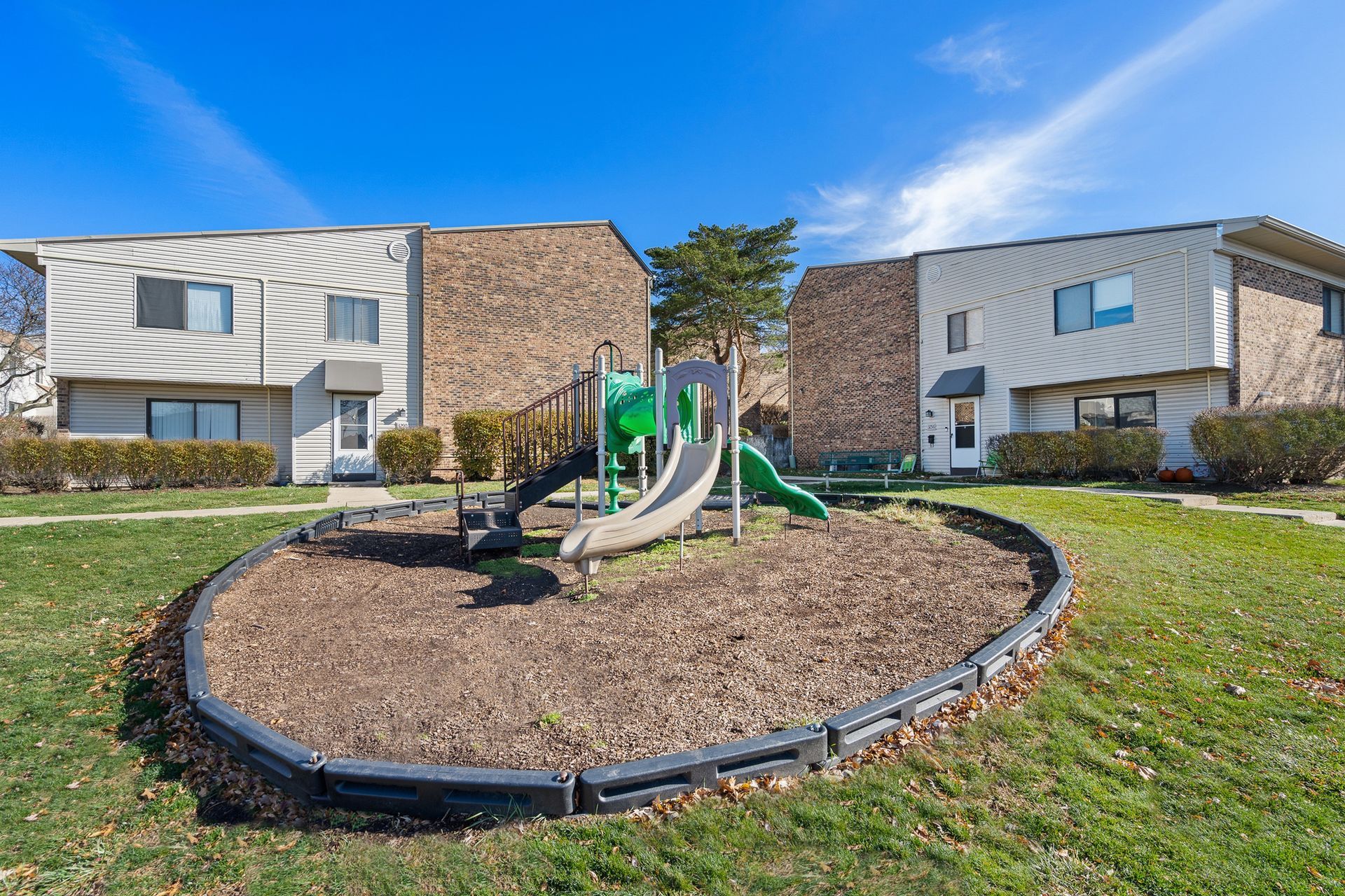 Playground with a slide in a mulch circle between two apartment buildings.