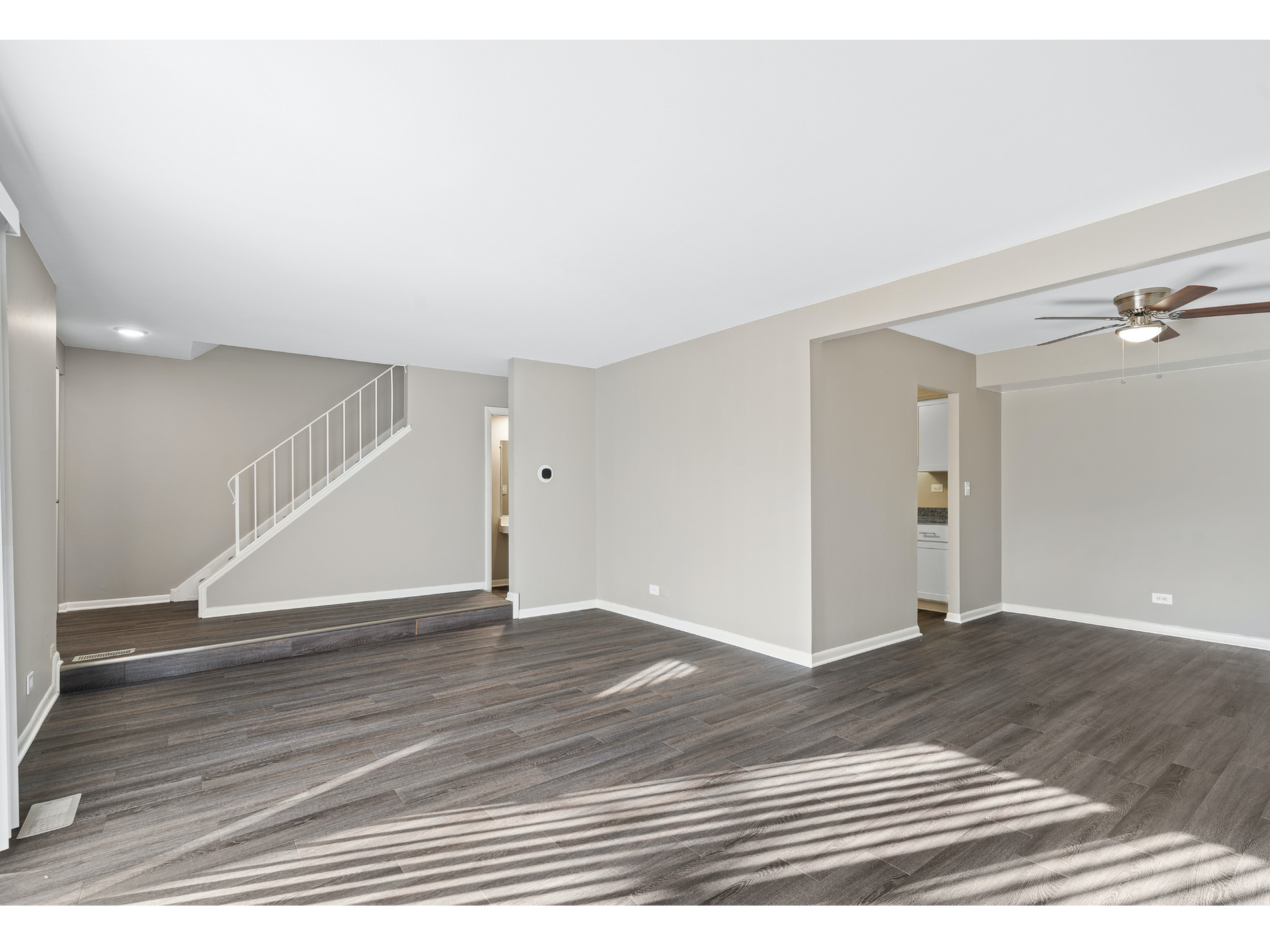 Bright living area with gray walls, dark wood-look flooring, staircase on the left, and a ceiling fan.