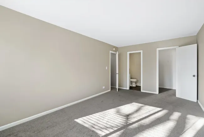 Empty room with gray carpet, beige walls, and white doors. Sunlight streams across the floor at Townhomes at Highcrest in Woodridge, IL.