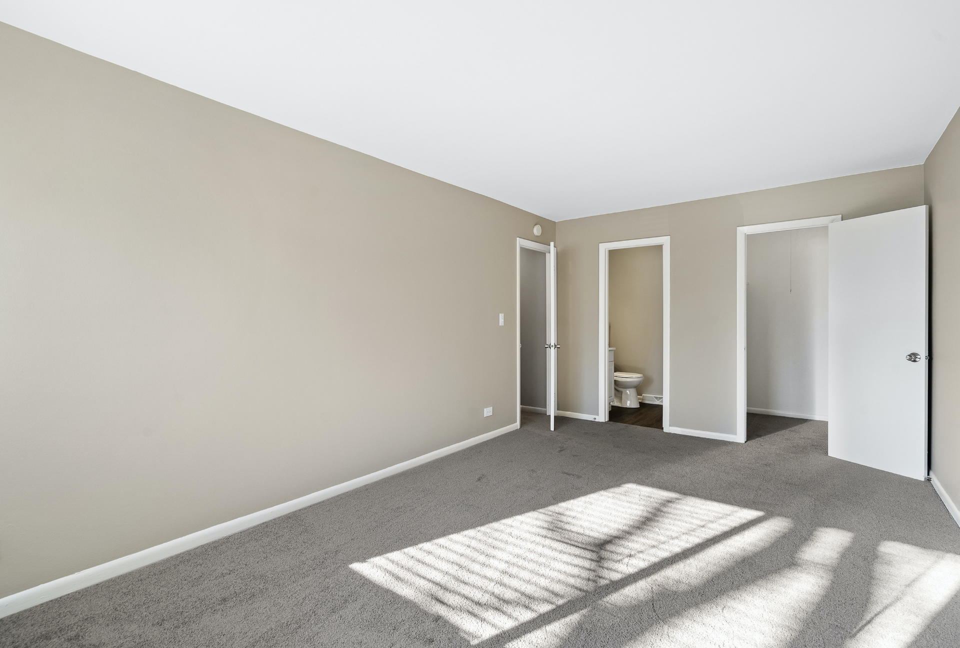 Empty beige bedroom with carpet, two open doors, and sunlight shadows on the floor.
