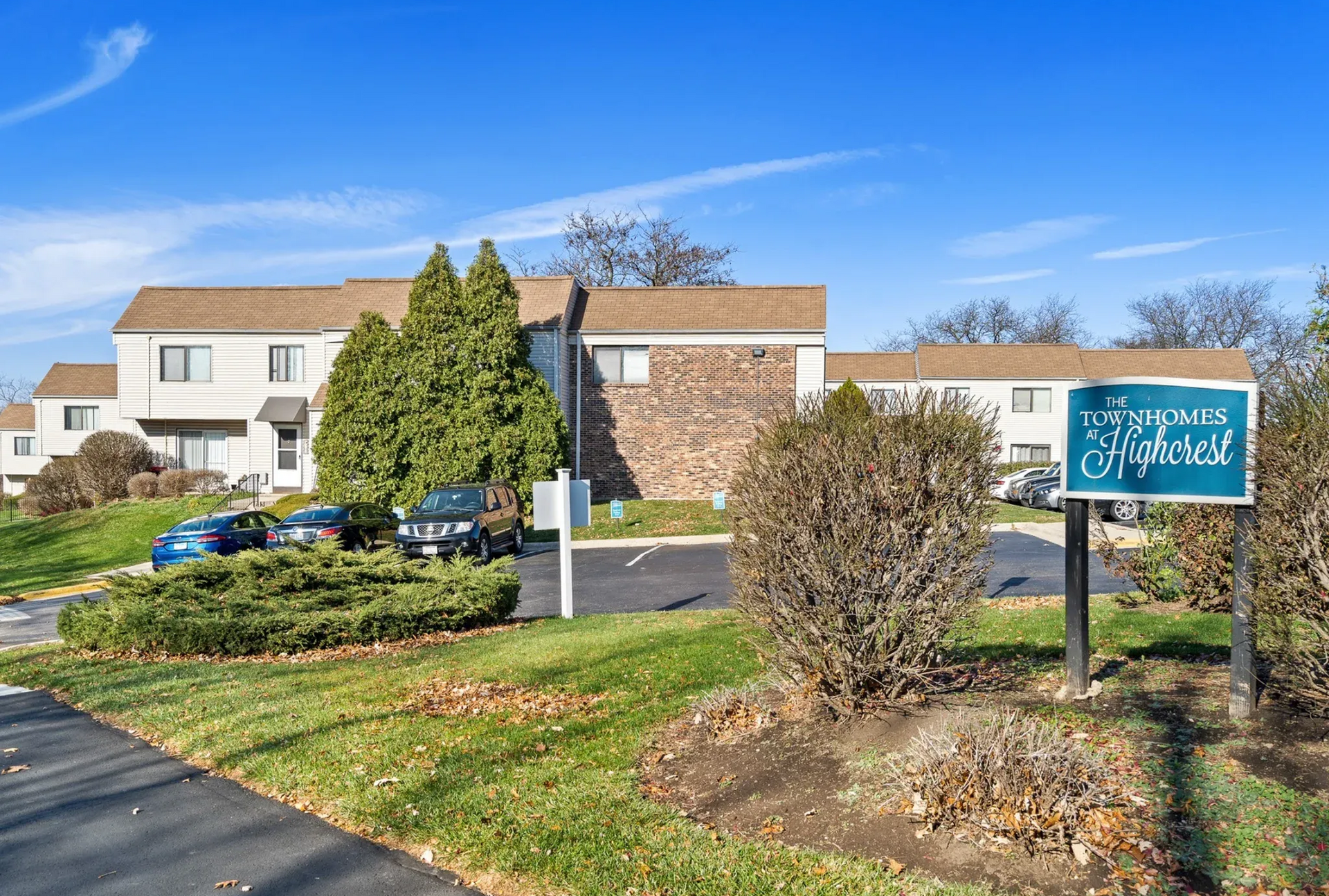 Exterior view of The Townhomes at Highcrest community with sign, cars, and landscaping.