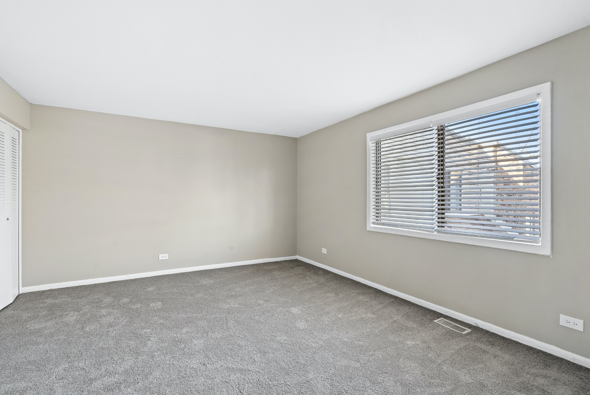 Empty beige bedroom with carpet, a window with white blinds, and a white closet door.