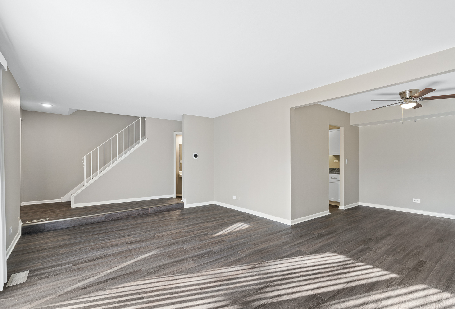 Bright living area with gray walls, dark wood-look flooring, staircase on the left, and a ceiling fan.